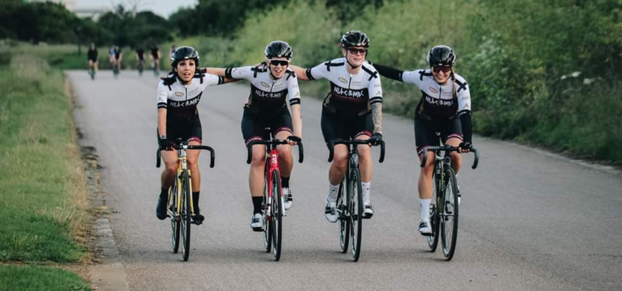 Caz and their team mates cycling outside down a round, surrounded by grass. They are holding the handlebars and have an arms on each other's shoulders whilst lined up alongside each other. 