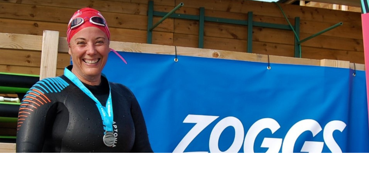 Alison smiling in her wesuit and swimming hat, wearing a medal in-front of a banner. 