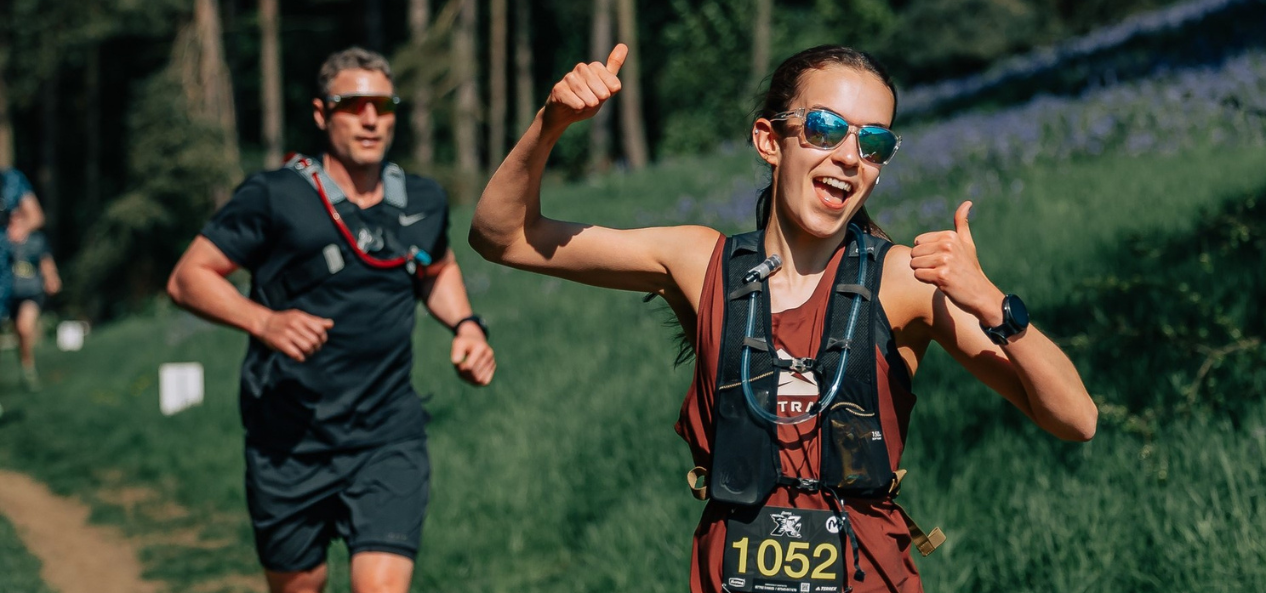 Elena smiling and trail running outside with another runner behind her. 