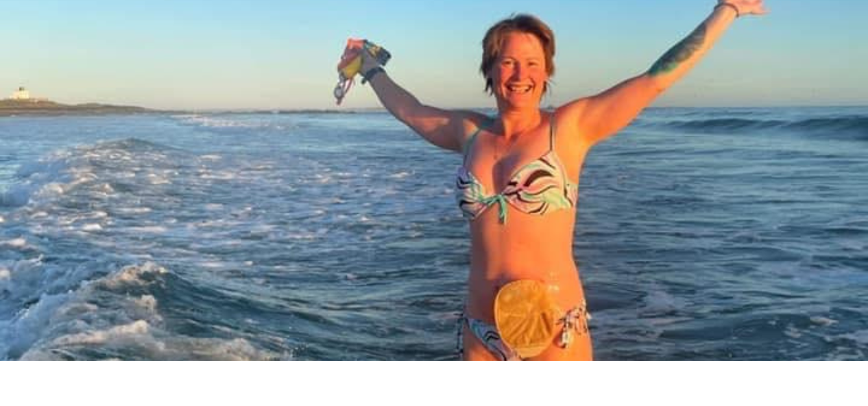 Gill wearing her bikini, smiling, with her arms raised in the arm in joy whilst standing in the sea. 