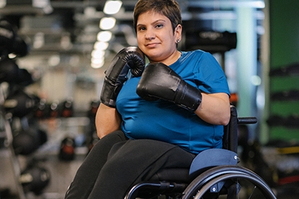 An Indian woman in a wheelchair poses for a photo in a gym, wearing boxing gloves.