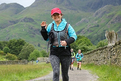 A woman waves to the camera while she walks in a forest. 