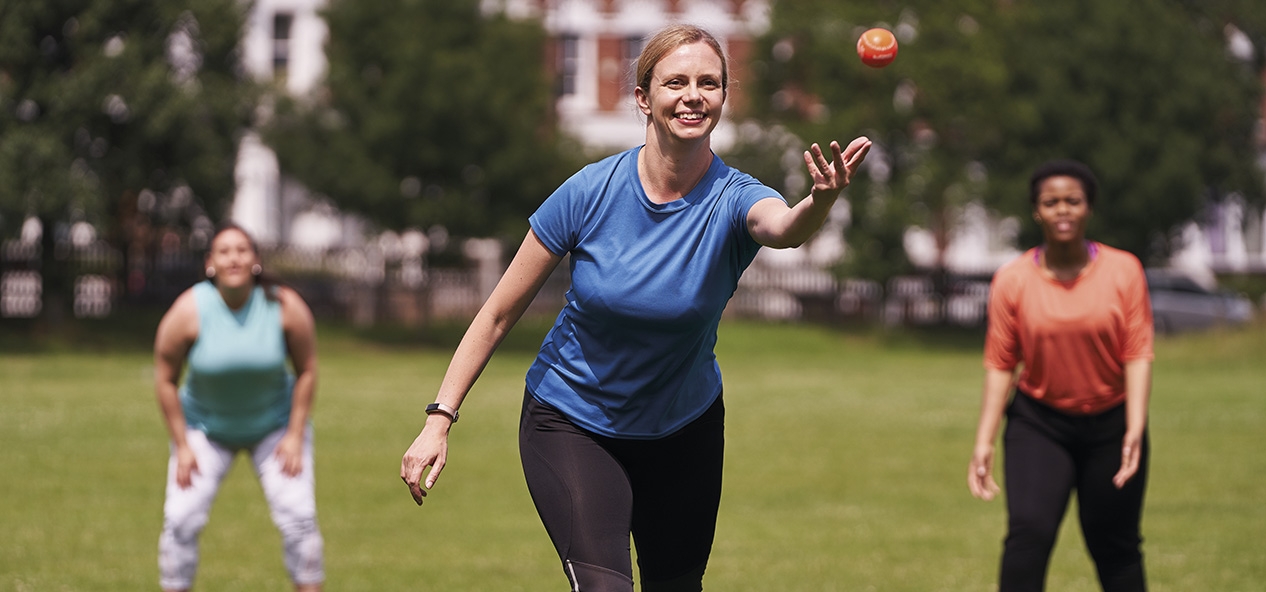 A woman throws a rounders ball as a group play in the park