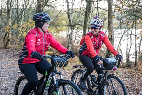 Two women ride bikes on a trail by a body of water