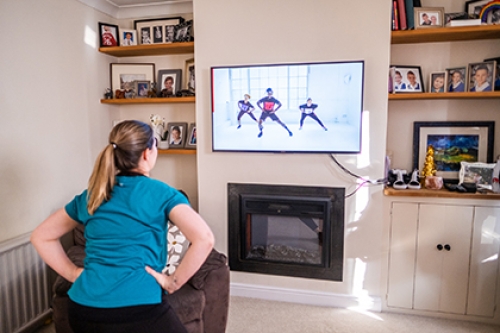 A woman exercises along with a Couch to Fitness session in her living room, watching the class on her TV