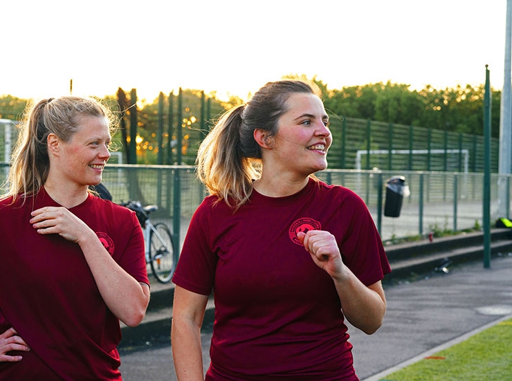 Two women stand at the side of a football pitch, about to play